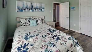 A bedroom with a floral bedspread, a large painting of sailboats, a white door, and a hallway leading to another room. - Fish Tales by Miss Kitty’s (Rockport)