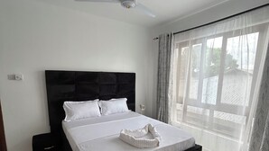 Ein Schlafzimmer mit einem großen Bett, einem Deckenventilator, einem Nachttisch, einem schwarzen Teppich und einem Fenster mit durchsichtigen Vorhängen.
