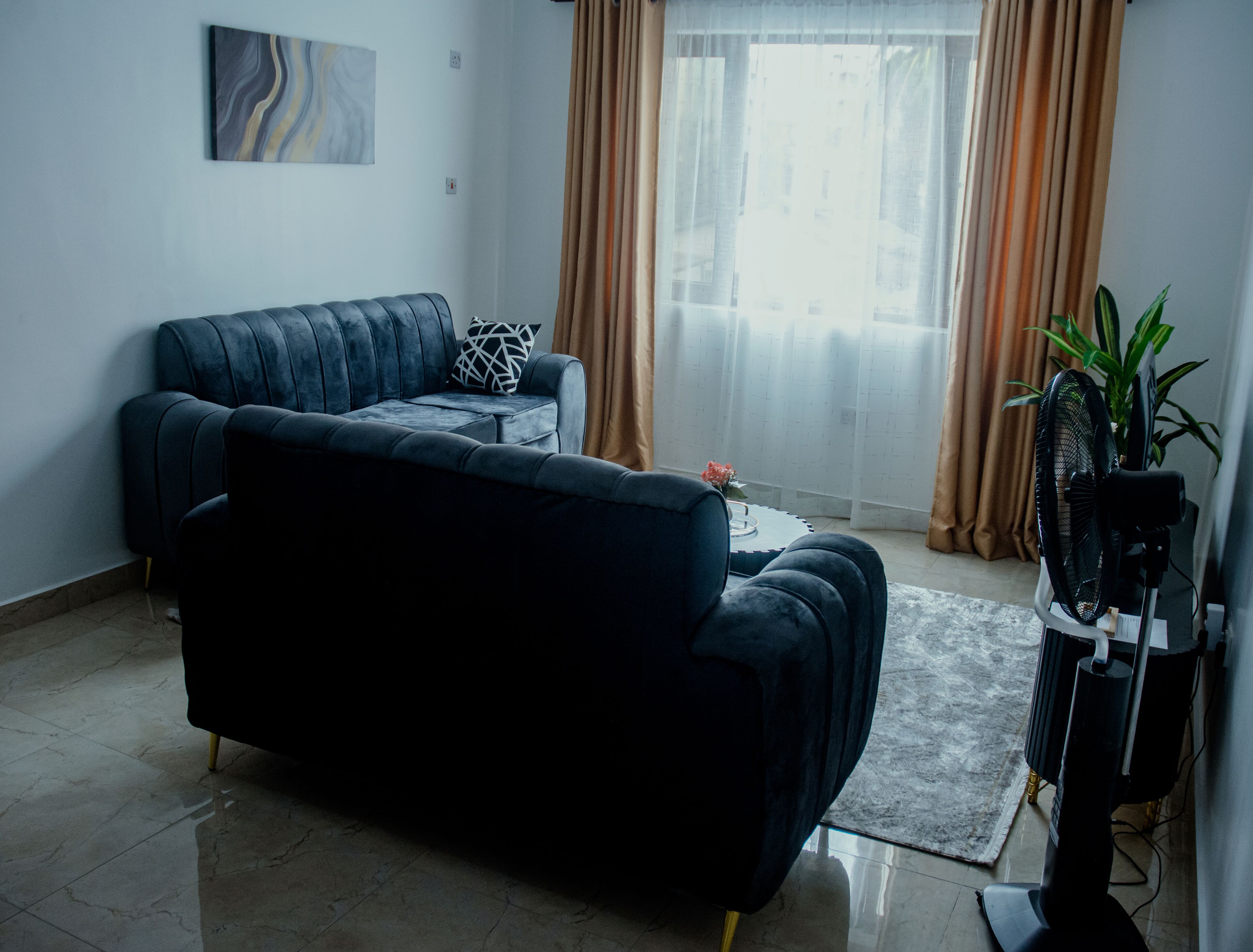 Un salon avec un canapé bleu foncé, une petite table d’appoint, une plante en pot et un meuble pour la télévision équipé d’un ventilateur.
