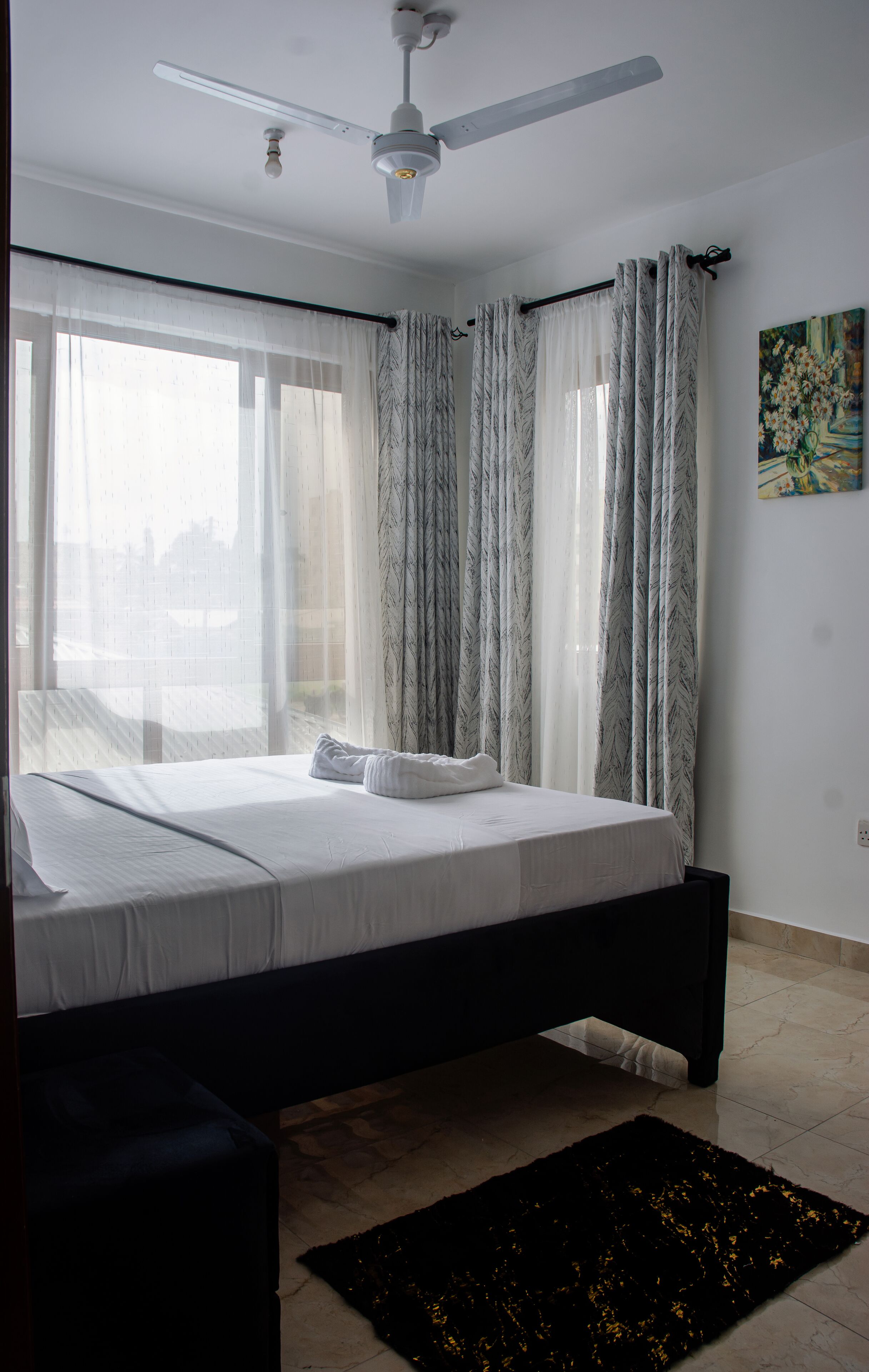 Un dormitorio con cama, ventilador de techo, ventana con cortinas traslúcidas y un cuadro en la pared.