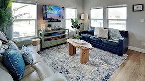 A living room with a beige sofa, a blue sofa, a coffee table, a TV on a stand, and a large window.