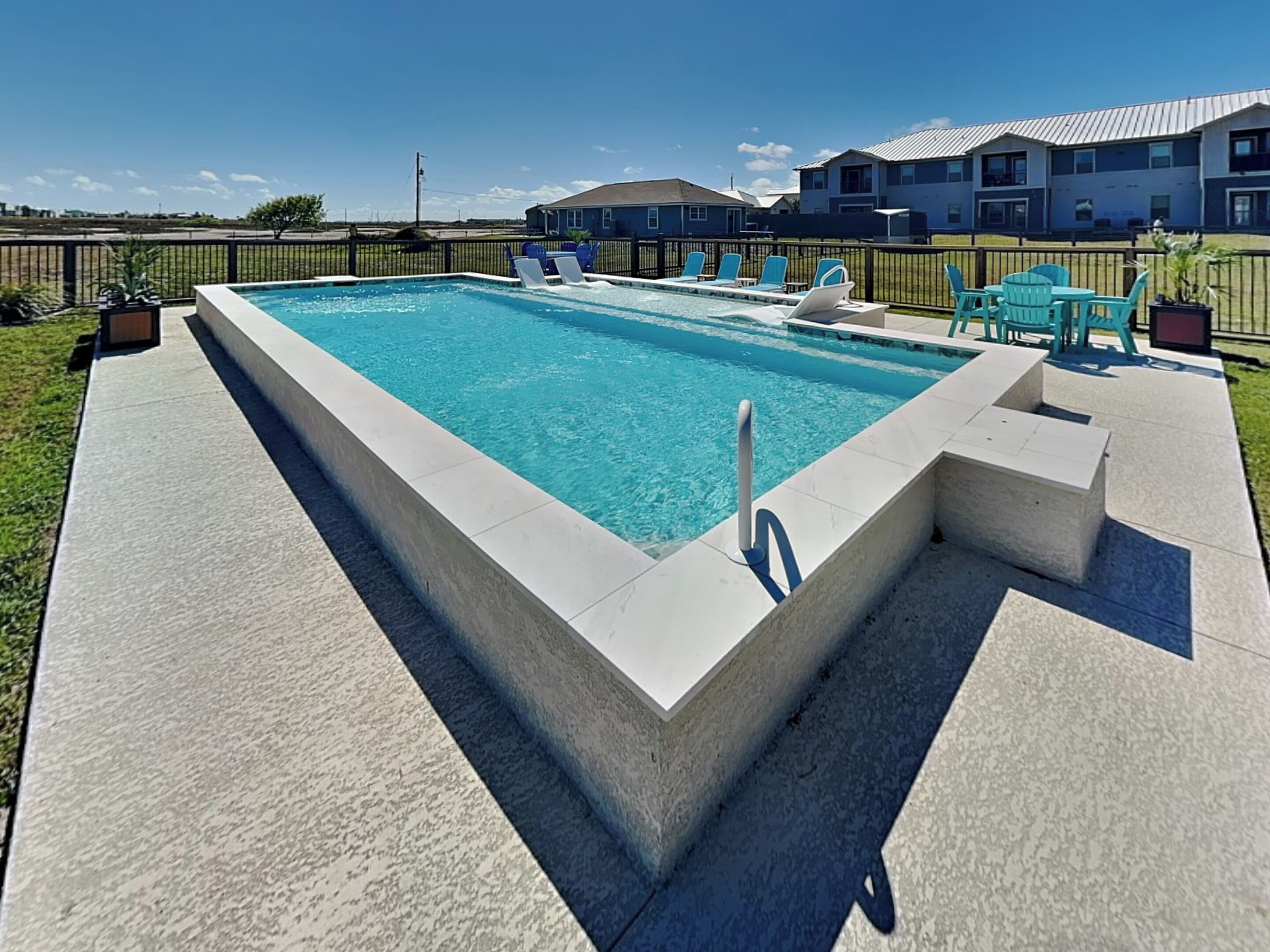 A rectangular swimming pool with a concrete deck, surrounded by a fence and a row of blue chairs.