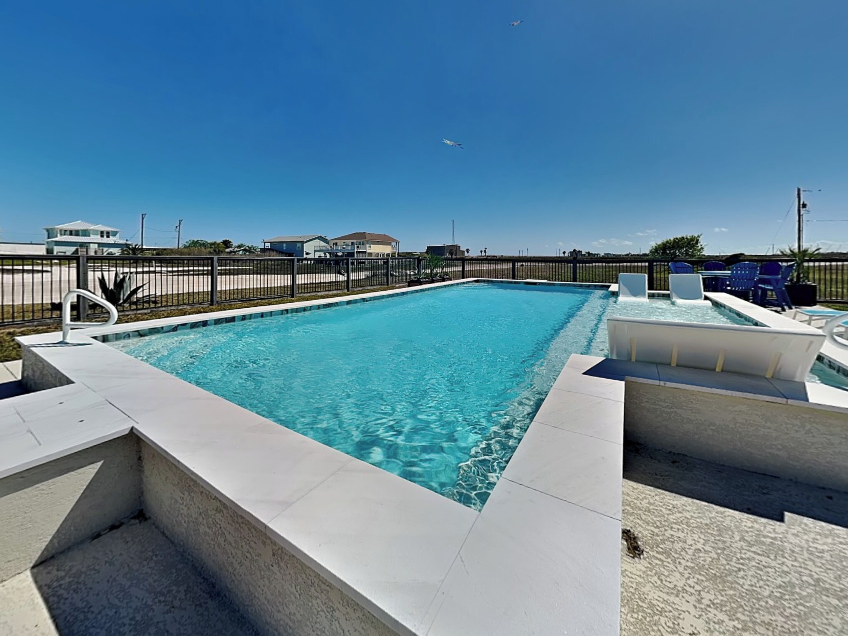 A rectangular swimming pool with clear blue water, surrounded by a black fence, and a white deck area with chairs and a table.