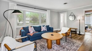 Ein modernes Wohnzimmer mit einem blauen Sofa, einem runden Holztisch und einem großen Fenster mit Vorhängen.
