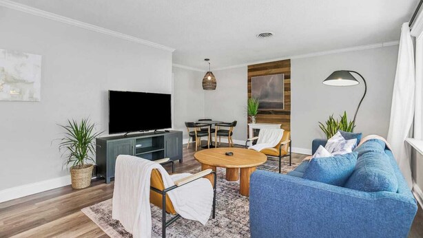 Una sala de estar moderna con un televisor de pantalla plana, una mesa redonda de madera, un sofá azul y un comedor con mesa y sillas de madera.