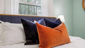 Ein Bett mit weißer Bettwäsche, einem dunkelblauen Kopfteil und mehreren Kissen in Schwarz, Blau und Orange.