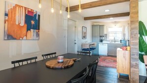 A dining area with a long table, chairs, and a wooden serving board. There is a painting on the wall and a kitchen in the background.