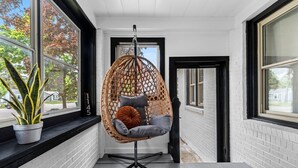 Una silla colgante con cojines en un pasillo con paredes de ladrillo blanco y ventanas grandes.