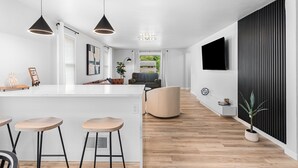 Una cocina moderna con una isla central, taburetes de bar, iluminación colgante y vistas a una sala de estar con sofá y televisor.