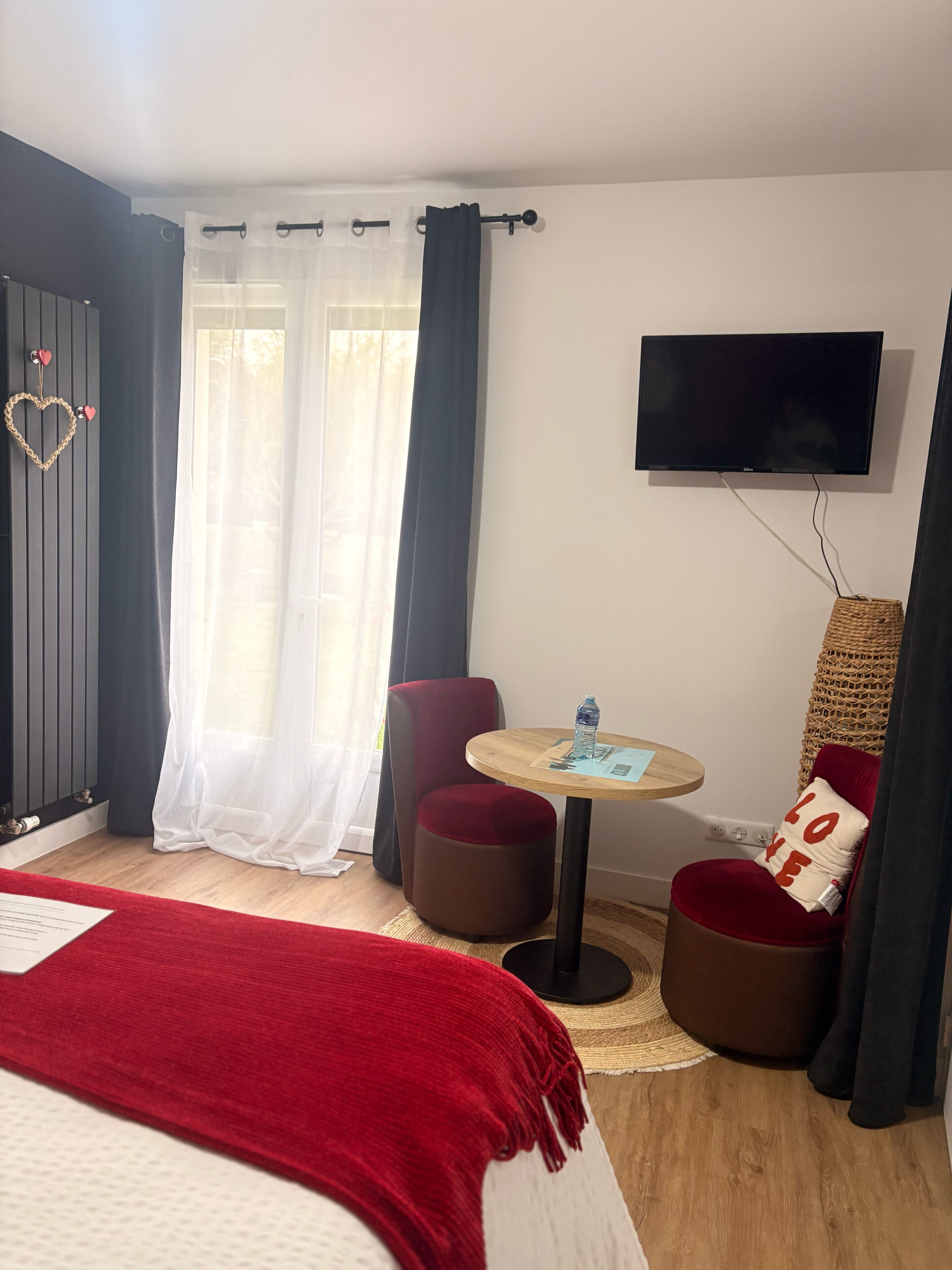 A hotel room with a bed, a round wooden table with a water bottle, a red chair, a television on the wall, and a window with sheer curtains.