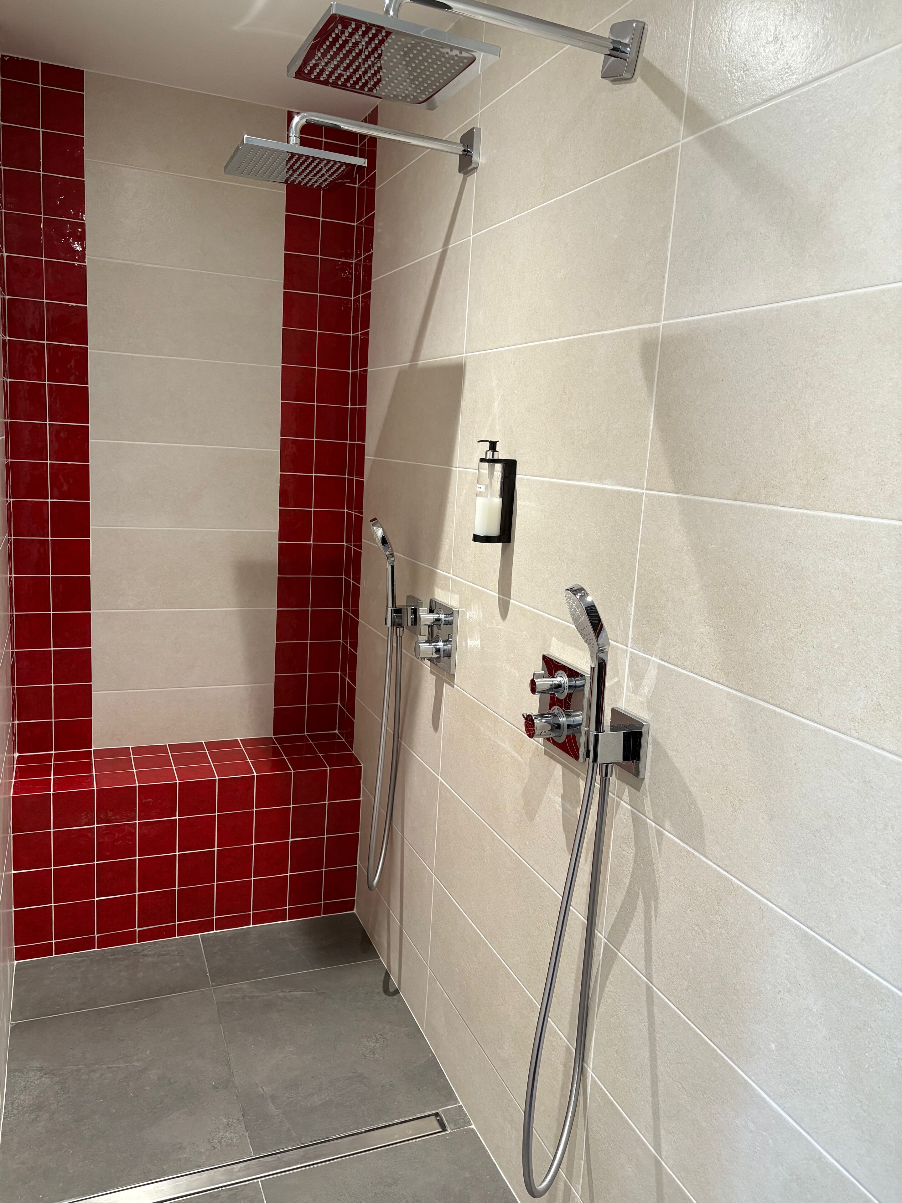 Zona de ducha con azulejos rojos y beige, un cabezal de ducha manual, un grifo empotrado y un dispensador de jabón.