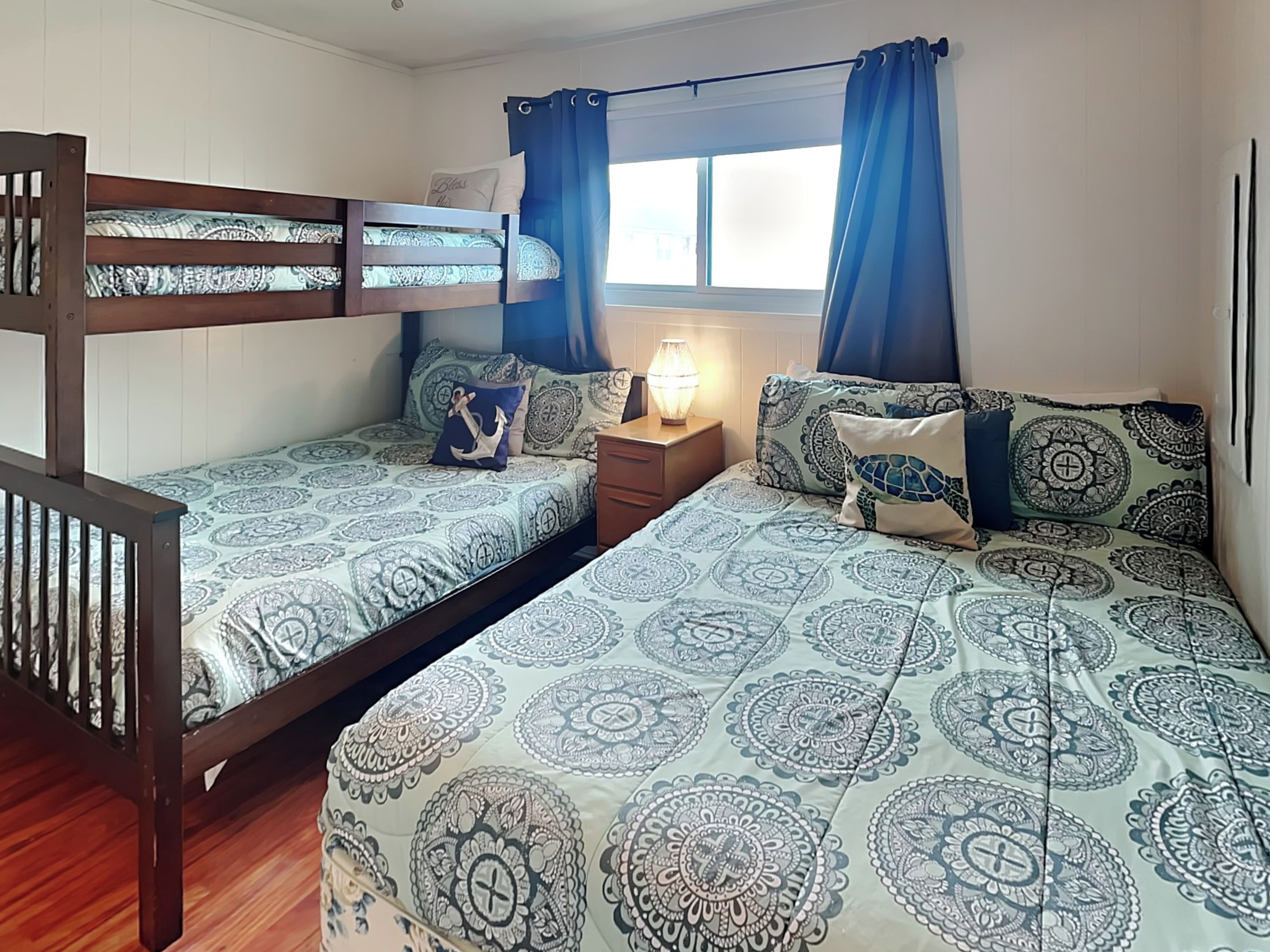Un dormitorio con una cama litera, dos camas individuales, ropa de cama estampada, una mesita de noche con lámpara y una ventana con cortinas.