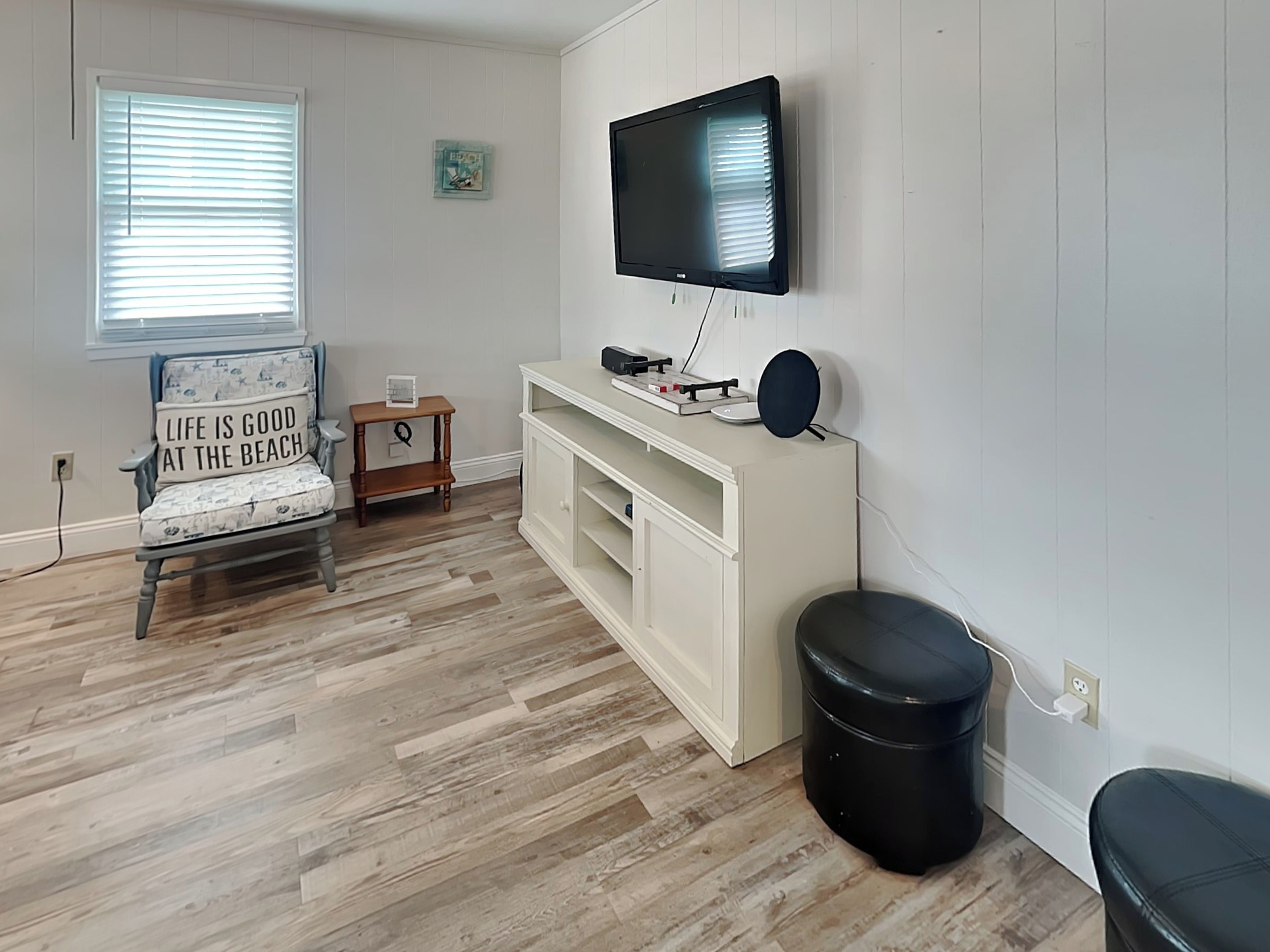 A room with a wooden floor, a white wall-mounted TV, a chair with a beach-themed cushion, a small wooden table, and a white cabinet.