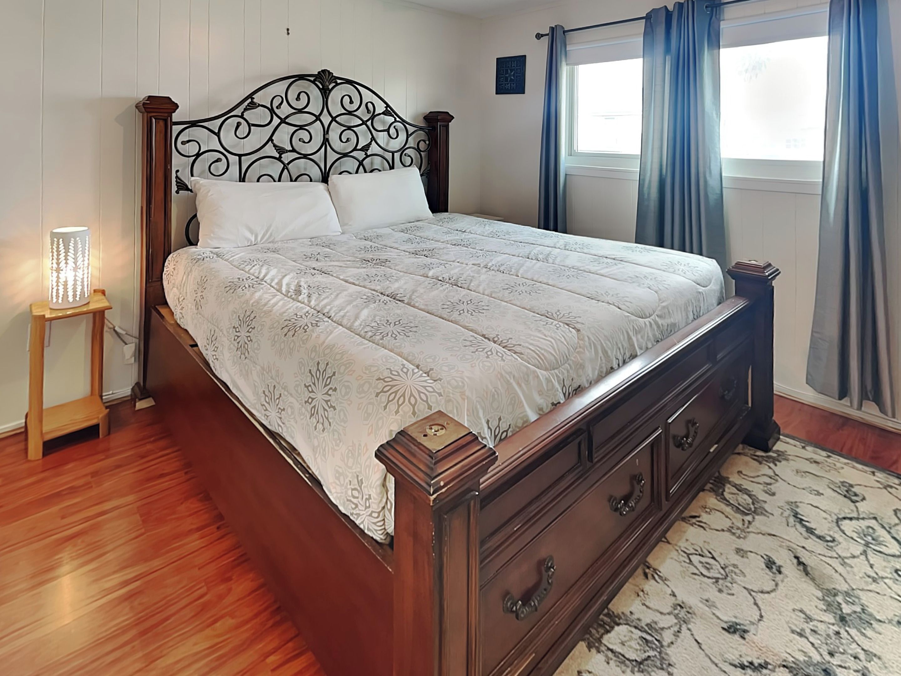Un dormitorio con estructura de cama de madera, colcha estampada, mesita de noche con lámpara y ventana con cortinas.