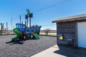 A playground with a slide and climbing structures, a small building with a garage door, and a gravel ground.