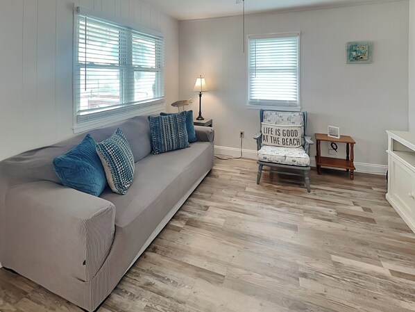 A living room with a grey sofa, blue cushions, a wooden chair with a beach-themed cushion, a small wooden table, and a lamp. - John Silver Cottage by Ryson Vacation Rentals (Galveston)