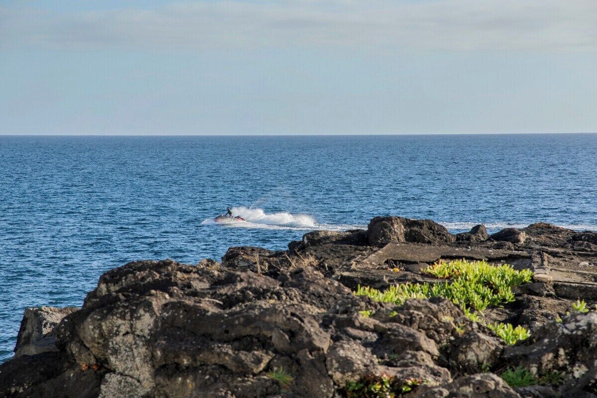 Une côte rocheuse bordée d’une eau d’un bleu limpide, avec un jet ski visible au loin.