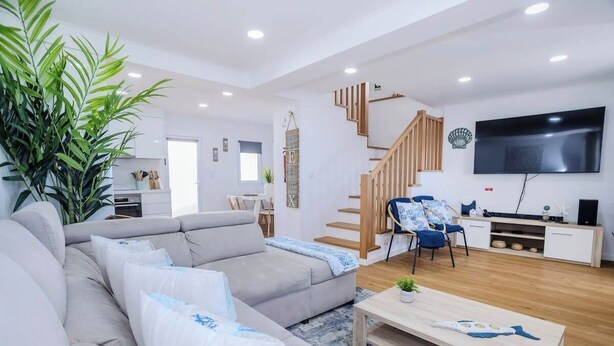 A modern living room with a sofa, coffee table, and a TV. There is a staircase with wooden railings and a dining area in the background. - GuestReady - Sea Escape Azores (Lagoa)