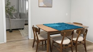 A dining area with a wooden table and four chairs, a blue cloth on the table, and a grey sofa in the background. - Modern 4 Bedroom with King bed (Lubbock)