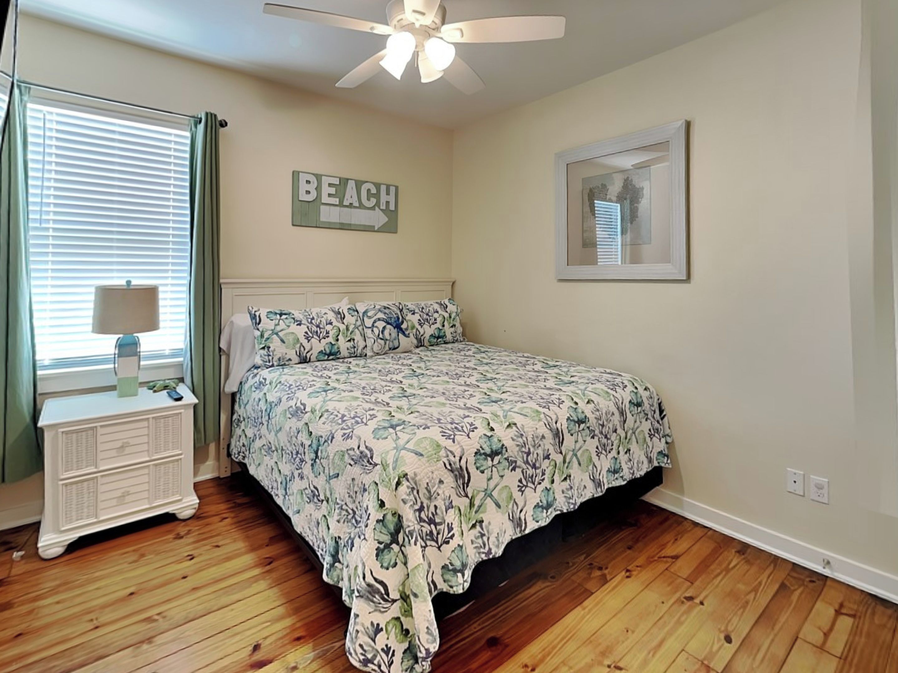 A bedroom with a bed, a nightstand, a lamp, a ceiling fan, a window with blinds, a wall sign reading "BEACH", and a framed picture.