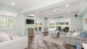 Ein geräumiges Wohnzimmer mit einer großen Couch, einem Couchtisch und einem an der Wand montierten Fernseher. Eine Glastür öffnet sich zu einem Außenbereich mit einem Pool.