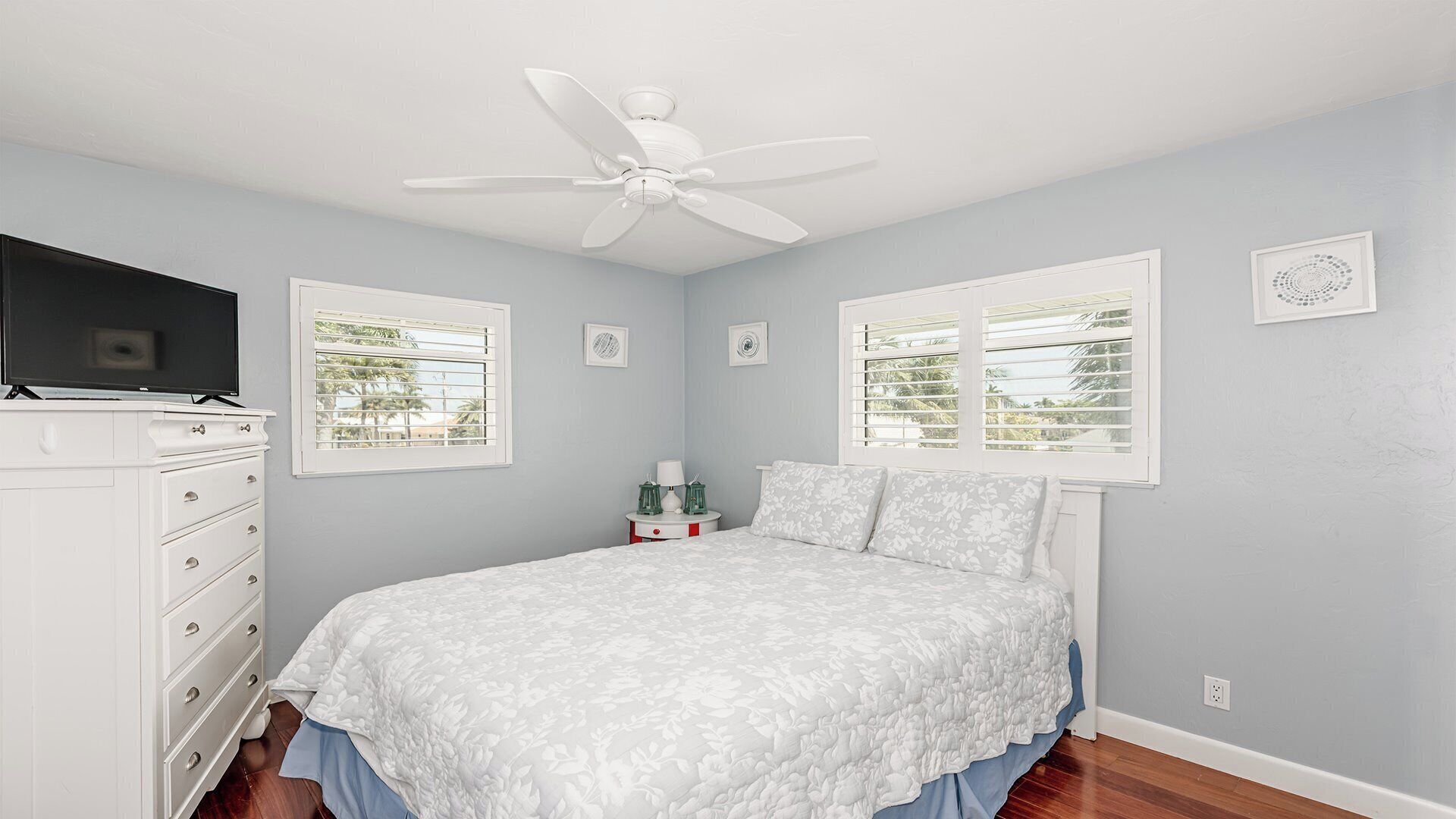 Une chambre à coucher comprenant un lit, une commode, un ventilateur de plafond et des fenêtres équipées de stores.