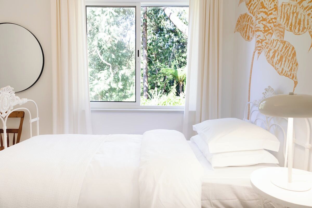 Une chambre avec une grande fenêtre, un lit recouvert de draps blancs, une table de chevet ronde et une lampe fixée au mur.