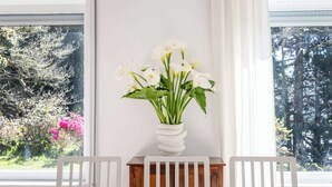 A dining area with a wooden table and white chairs, a white vase with white flowers, and a view of trees and shrubs through large windows.