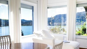 A room with a wooden dining table, white chairs, a white sofa, and large windows offering a view of a lake and mountains.