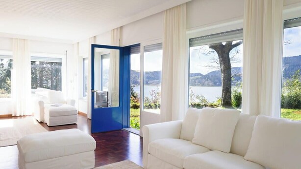 A modern living room with a white sofa, a white armchair, and a matching ottoman. A large window offers a view of a lake and trees.