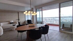 A modern living room with a round dining table, black chairs, a white sofa, and a large window offering a view of a marina.