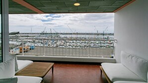 A balcony with a view of a marina and construction cranes. There are two white sofas and a wooden table.