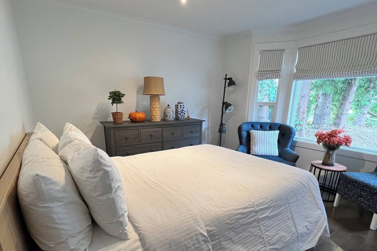 A modern bedroom with a large bed, a dresser, a chair, and a window with a view of trees.