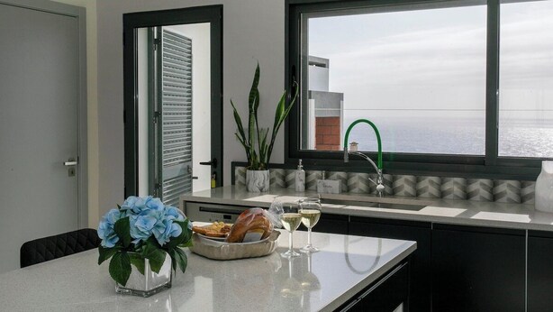 A modern kitchen with a view of the sea, a dining table with a vase of flowers, and a buffet with bread and wine glasses. - GuestReady - Sunset House (Relva)