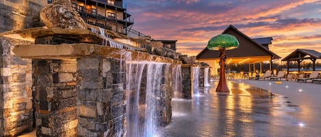 A stone waterfall feature with a wooden deck and lounge chairs, a building with balconies, and a sunset sky.