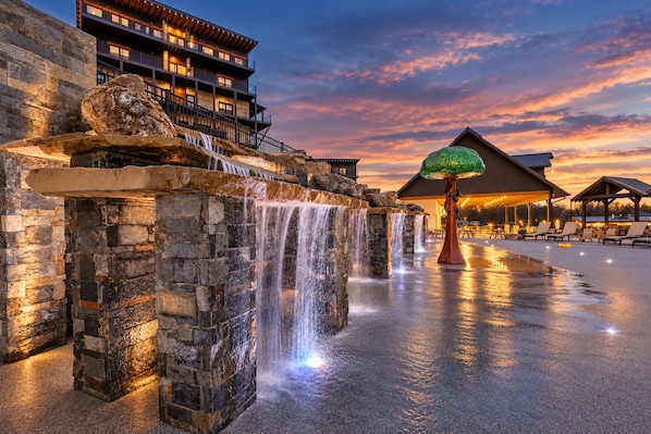 A stone waterfall feature with a wooden deck and lounge chairs, a building with balconies, and a sunset sky.