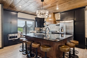 A modern kitchen with dark cabinetry, a central island with stools, stainless steel appliances, and a wooden ceiling. - Indoor Pool | Rooftop Lounge | Theater | Sleeps 22 | Free Waterpark (Sevierville)