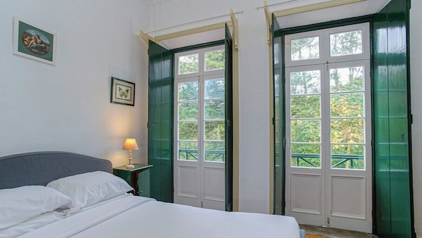 A bedroom with a bed, a nightstand with a lamp, two green-framed doors, and a window with a view of greenery.
