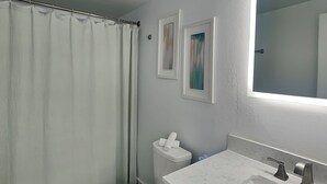 A bathroom with a shower curtain, toilet, sink, and framed artwork on the wall. - The Wave Cave by Silver Sands Vacation Rentals (Port Aransas)