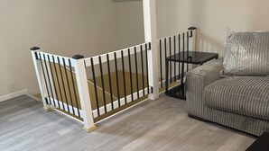 Un cuarto estilo loft con piso de madera, barandal blanco, sofá gris y una pequeña mesa negra.