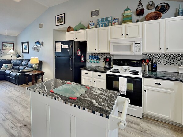 A modern kitchen with white cabinets, a black refrigerator, a stove, and a microwave. There is a dining area with a black leather couch and a small table with a lamp. - Southern Comfort by Silver Sands Vacation Rentals (Port Aransas)