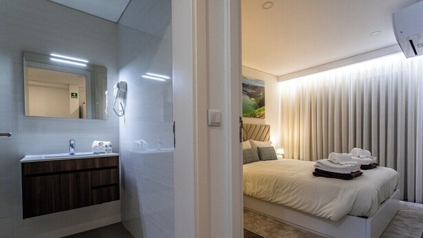 A modern bathroom with a sink, mirror, and towel warmer. The bedroom features a bed with neatly folded towels and a large window with blinds. - GuestReady - GuestHouse Passal - S. Miguel (Ponta Delgada)