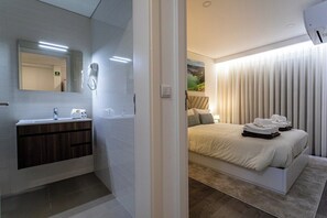 Een moderne badkamer met een wastafel, spiegel en handdoekwarmer. De slaapkamer heeft een bed met netjes opgevouwen handdoeken en een groot raam met luxaflex.