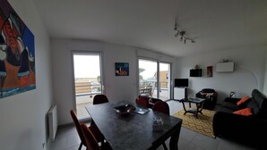 Ein geräumiger Wohnbereich mit Esstisch, Stühlen, einer Couch und einem Fernseher. Große Fenster und ein Balkon sind vorhanden.