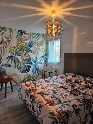 Une chambre avec une couvre-lit à motifs floraux, un fauteuil rouge, un mur recouvert d’un papier peint à motifs de feuilles tropicales et un luminaire suspendu.