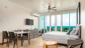 Een slaapkamer met een bed, een bureau, stoelen, een televisie en een balkon met uitzicht op zee.