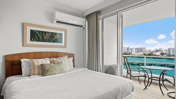 Habitación de hotel con cama, un cuadro enmarcado, unidad de aire acondicionado, balcón con mesa y sillas, y vistas a la ciudad y al agua.