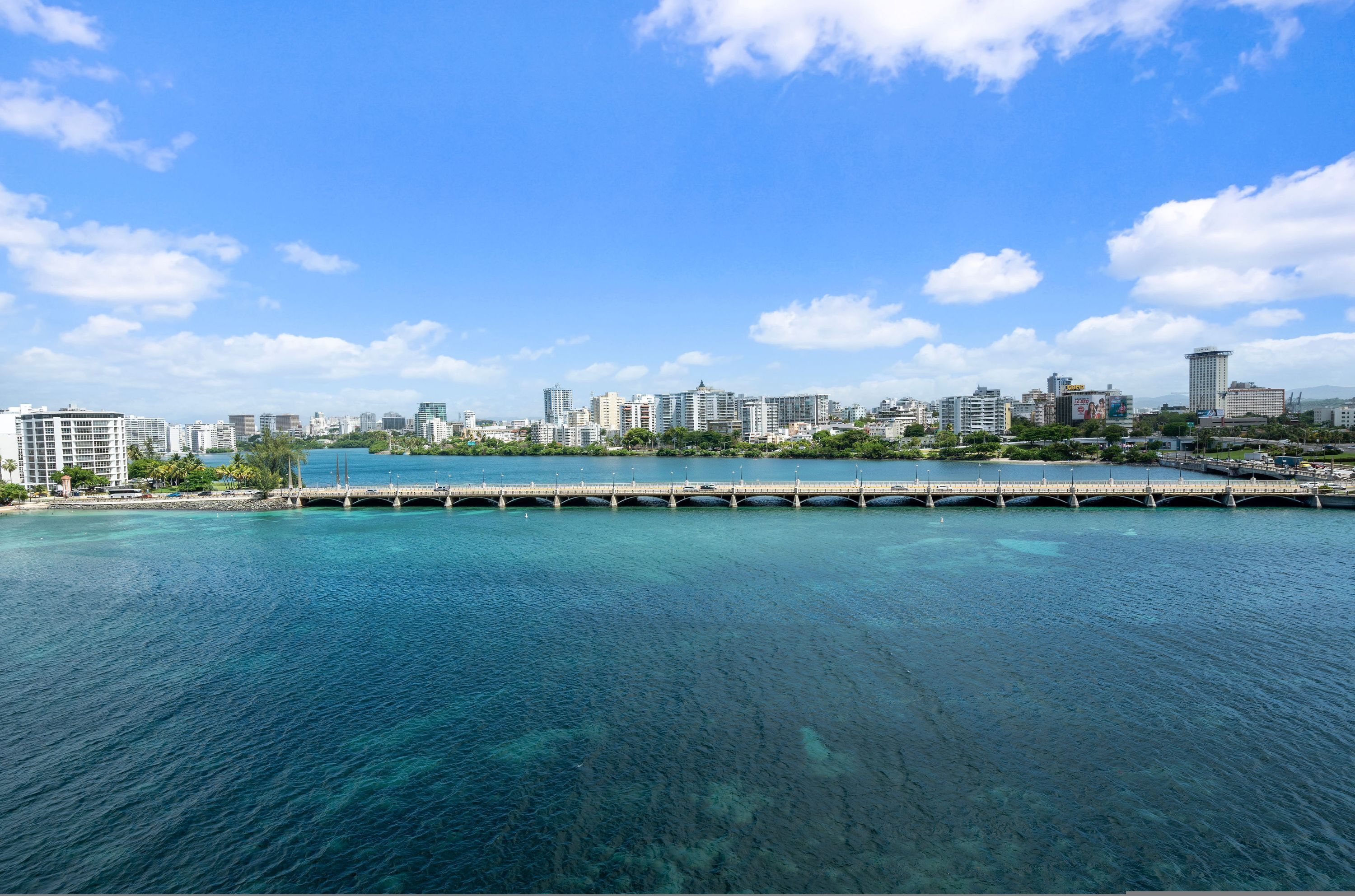 Un paisaje urbano costero con un puente destacado sobre aguas azul cristalino, edificios altos y un cielo despejado.