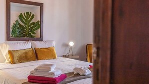 Un lit bien fait, avec des serviettes blanches et roses, une plante décorative accrochée au mur et une table de chevet avec une lampe.