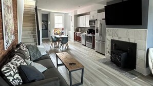 A modern living room with a sofa, coffee table, and fireplace. A kitchen with appliances and a dining area with chairs is visible.